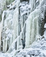 A person dressed in winter clothing stands next to a massive frozen waterfall. The long icicles cascade down a rocky cliff, surrounded by snow-covered terrain and trees peeking through. The setting appears serene and awe-inspiring, with the person gazing up at the natural ice formations.