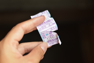 A hand holding torn pieces of a ticket with numbers and Thai writing on it. The background is blurred and dark, making the pieces in the foreground stand out.