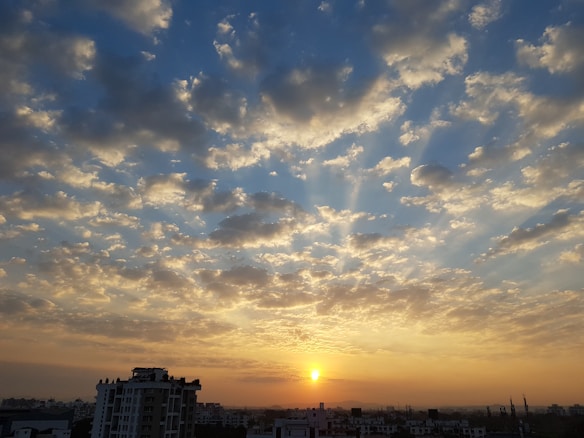 A vibrant sunrise over an urban skyline where the sun is partially visible on the horizon. The sky is filled with numerous scattered clouds, creating a beautiful pattern with varying shades of blue, orange, and gold. Tall buildings are silhouetted against the morning light at the bottom of the image.