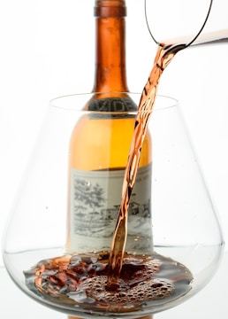 A close-up view of a glass being filled with a dark, amber-colored liquid, possibly wine or a spirit. In the background, an amber bottle with a vintage-style label is visible, suggesting a refined setting.