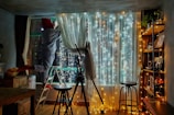 A cozy indoor setting with string lights illuminating a room. A person wearing red socks is standing on a ladder, adjusting curtains in front of a window. Shelves lined with books and decorative items, along with bar stools, create a warm and inviting atmosphere.