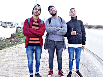 Three young men stand confidently on a paved pathway. They are wearing casual clothing, including jeans, jackets, and sneakers. The background shows a suburban area with grass and distant buildings.