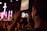 A person is holding a smartphone to capture a live event, focusing on a stage with performers. The stage is illuminated with purple and pink lights, and a large cross is visible in the background. The scene appears to be in a dimly lit auditorium or venue filled with attendees.