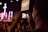 A person is holding a smartphone to capture a live event, focusing on a stage with performers. The stage is illuminated with purple and pink lights, and a large cross is visible in the background. The scene appears to be in a dimly lit auditorium or venue filled with attendees.