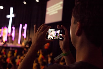 A person is holding a smartphone to capture a live event, focusing on a stage with performers. The stage is illuminated with purple and pink lights, and a large cross is visible in the background. The scene appears to be in a dimly lit auditorium or venue filled with attendees.