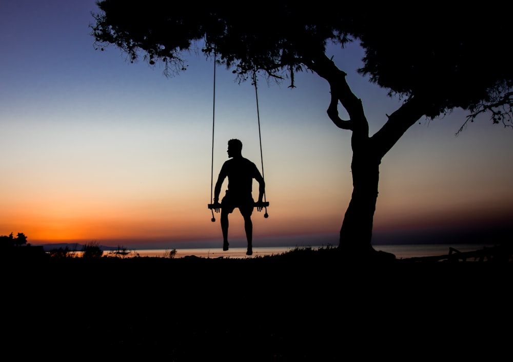 silhouette photo of man on swing beside tree
