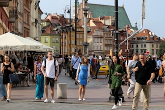 A bustling street scene in a historic European city, with people walking and interacting. The background features colorful old buildings with a variety of architectural styles and a church with a prominent green roof. An outdoor seating area with a canopy labeled 'CIECHAN' is visible, along with a yellow vehicle.