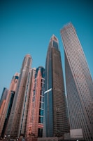 A cluster of modern skyscrapers with varying architectural designs reaches toward the clear blue sky. The buildings display glass facades and a mix of straight and curved lines, showcasing urban development and innovation.