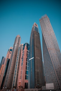 A cluster of modern skyscrapers with varying architectural designs reaches toward the clear blue sky. The buildings display glass facades and a mix of straight and curved lines, showcasing urban development and innovation.