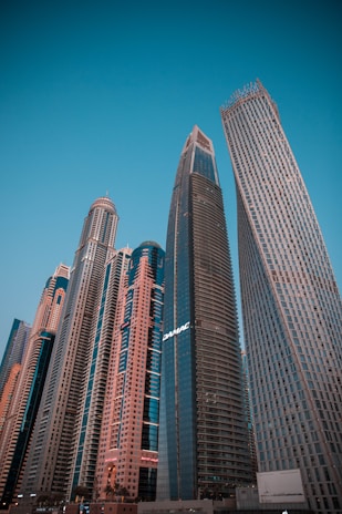 A cluster of modern skyscrapers with varying architectural designs reaches toward the clear blue sky. The buildings display glass facades and a mix of straight and curved lines, showcasing urban development and innovation.