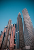 A cluster of modern skyscrapers with varying architectural designs reaches toward the clear blue sky. The buildings display glass facades and a mix of straight and curved lines, showcasing urban development and innovation.
