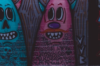 A vibrant graffiti artwork on a brick wall featuring cartoonish figures with exaggerated facial features. The designs are colorful and incorporate various patterns, with elements like big eyes, large teeth, and whimsical facial expressions.
