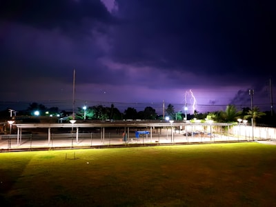 A dark night sky is illuminated by a vivid lightning strike near a backdrop of silhouetted trees and power lines. In the foreground, a fenced area with a carport or shelter is visible, highlighted by artificial lighting. The grass appears neatly mowed and stretches out towards the street lamps lining the unseen path.