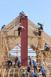 Several individuals in blue shirts and straw hats are constructing a wooden building framework. They are manually installing red panels onto the structure under a clear blue sky.