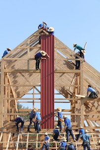 Several individuals in blue shirts and straw hats are constructing a wooden building framework. They are manually installing red panels onto the structure under a clear blue sky.