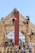 Several individuals in blue shirts and straw hats are constructing a wooden building framework. They are manually installing red panels onto the structure under a clear blue sky.