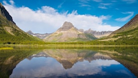 A serene mountain landscape featuring a clear, reflective lake in the foreground. The lake reflects the surrounding mountains and bright blue sky, creating a mirror-like effect. Vegetation covers parts of the mountains, with dense green forests and patches of snow visible on the peaks.