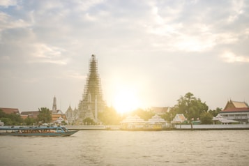 A majestic temple structure, partially under renovation, stands prominently on the riverbank against a bright sunset. Surrounding the temple are lush green trees and other traditional buildings, reflecting intricate architectural designs. A boat with vibrant blue accents is gliding across the river in the foreground.