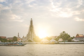 A majestic temple structure, partially under renovation, stands prominently on the riverbank against a bright sunset. Surrounding the temple are lush green trees and other traditional buildings, reflecting intricate architectural designs. A boat with vibrant blue accents is gliding across the river in the foreground.