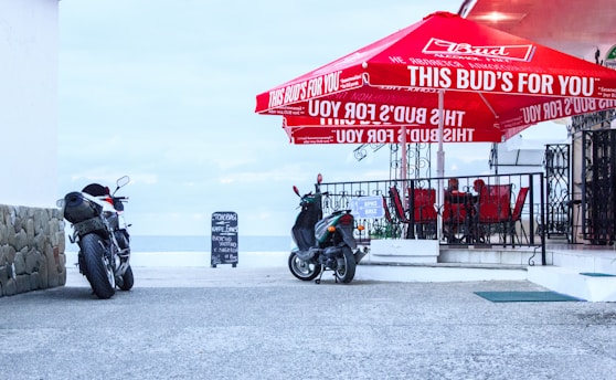 A seaside cafe with a red Budweiser branded umbrella over a set of outdoor tables and chairs. Two motorcycles are parked on the pavement nearby. A chalkboard sign stands outside the cafe, with sea and sky visible in the background.