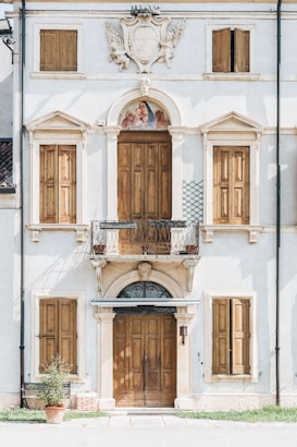 A classic European-style building facade with wooden shutters covering the windows. Above the central wooden door is a small balcony with ornate metalwork. A decorative stone relief with a shield and foliage motifs is prominently displayed at the top, and a religious fresco featuring several figures is set into an arch above the balcony.