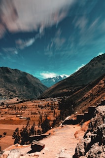 A breathtaking mountainous landscape features a winding path bordered by rocks, leading toward a horizon with snow-capped peaks. The sky displays dramatic, fast-moving clouds against a vibrant blue backdrop. The valley below is dotted with fields and sparse trees, creating a serene yet majestic scene.