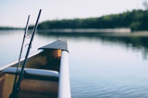 A calm river scene is depicted with the front of a canoe visible in the foreground. Two fishing rods are mounted on the canoe, hinting at a peaceful fishing trip. The background features a tranquil body of water reflecting the surrounding greenery and a bright sky.