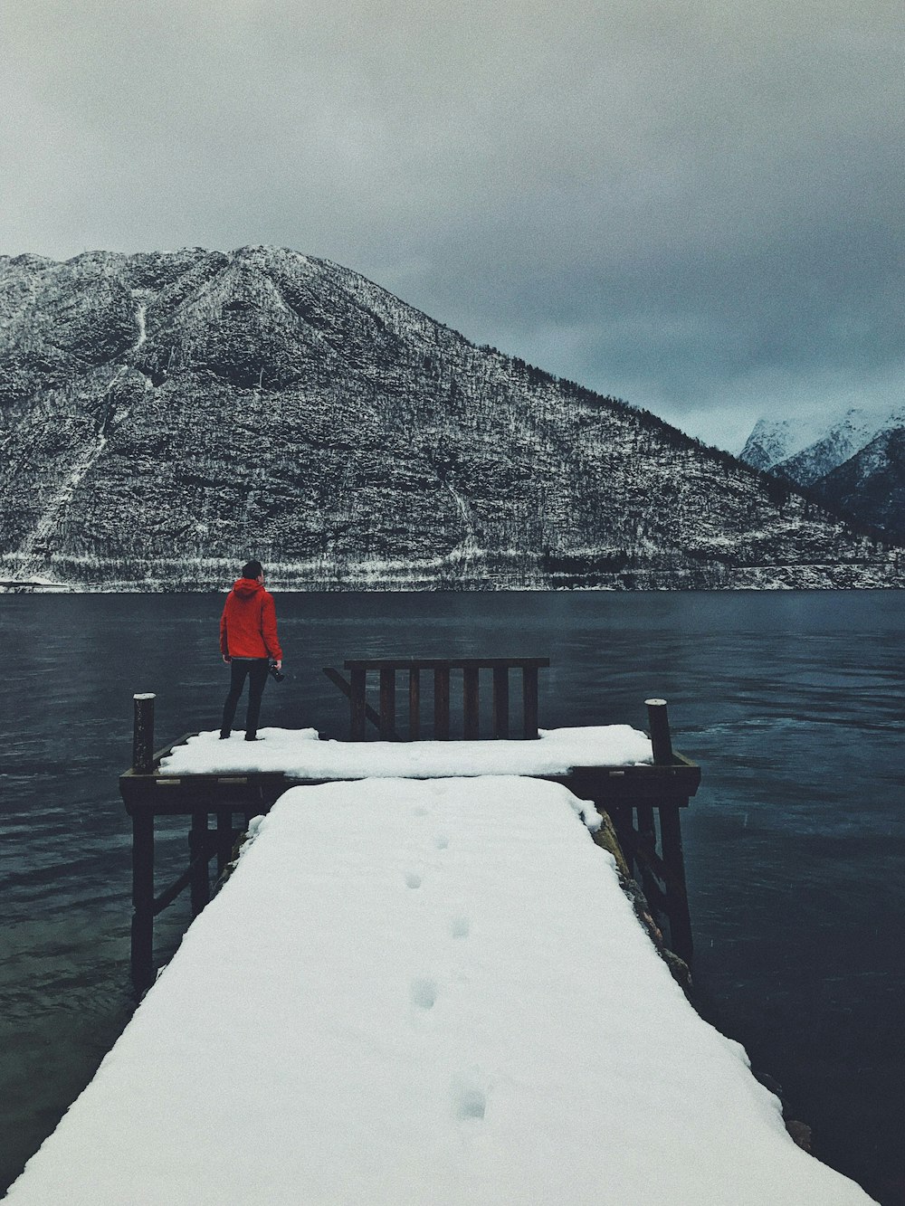 person standing on dock covered with snow looking at mountain across body of water