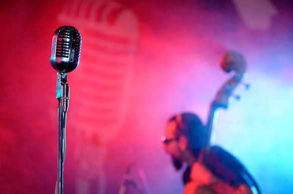 A vintage-style microphone is prominently featured in the foreground, sharply in focus against a vibrant, colorful background. The background shows a musician holding an upright bass, blurred, with colorful red and blue lighting creating a dynamic stage atmosphere.