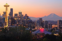 A cityscape during sunset showcasing tall buildings with illuminated windows and the iconic Space Needle on the left side. In the background, a mountain with a snow-capped peak is visible. The sky displays gradients of orange, pink, and purple hues. The overall scene includes a blend of natural and urban elements.