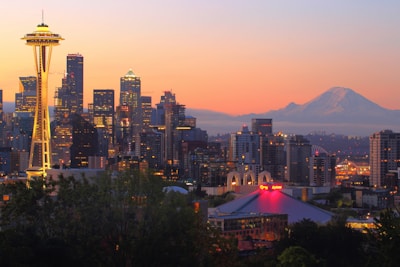 A cityscape during sunset showcasing tall buildings with illuminated windows and the iconic Space Needle on the left side. In the background, a mountain with a snow-capped peak is visible. The sky displays gradients of orange, pink, and purple hues. The overall scene includes a blend of natural and urban elements.