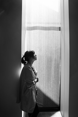 A person stands in a narrow, dimly lit space, gazing upwards towards a source of light filtering through a sheer curtain. The individual is dressed in a jacket and is leaning against the wall, conveying a sense of introspection or contemplation.