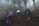 A group of hikers is trekking up a wooded hillside surrounded by dense fog. The path is muddy and uneven, with rough wooden railings on the sides. Tall trees with sparse foliage are visible, enhancing the misty atmosphere.