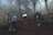 A group of hikers is trekking up a wooded hillside surrounded by dense fog. The path is muddy and uneven, with rough wooden railings on the sides. Tall trees with sparse foliage are visible, enhancing the misty atmosphere.
