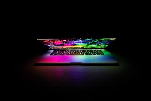 A laptop partially closed, emitting a vibrant spectrum of colors across its keyboard and screen. The colorful light is set against a dark, shadowy background, highlighting the brilliance of the hues.