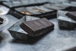 A close-up view of a chocolate bar partially removed from its mold, resting on a metallic surface. The background includes several other molds indicating a setting that resembles a kitchen or workspace for chocolate making. The surface reflects light, and the rich dark color of the chocolate contrasts with the dull silver of the molds.