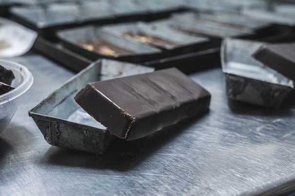 A close-up view of a chocolate bar partially removed from its mold, resting on a metallic surface. The background includes several other molds indicating a setting that resembles a kitchen or workspace for chocolate making. The surface reflects light, and the rich dark color of the chocolate contrasts with the dull silver of the molds.