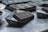 A close-up view of a chocolate bar partially removed from its mold, resting on a metallic surface. The background includes several other molds indicating a setting that resembles a kitchen or workspace for chocolate making. The surface reflects light, and the rich dark color of the chocolate contrasts with the dull silver of the molds.