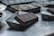 A close-up view of a chocolate bar partially removed from its mold, resting on a metallic surface. The background includes several other molds indicating a setting that resembles a kitchen or workspace for chocolate making. The surface reflects light, and the rich dark color of the chocolate contrasts with the dull silver of the molds.