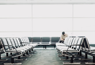 The scene depicts an empty waiting area with rows of black leather seats. A single person is seated in the back row, engrossed in an activity, possibly reading or using a device. The large glass windows in the background allow ample natural light to illuminate the area, creating a clean and spacious environment.