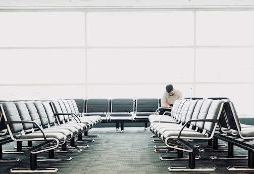 The scene depicts an empty waiting area with rows of black leather seats. A single person is seated in the back row, engrossed in an activity, possibly reading or using a device. The large glass windows in the background allow ample natural light to illuminate the area, creating a clean and spacious environment.