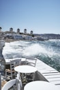 A coastal scene with several traditional windmills lined up on a hillside overlooking the sea. In the foreground, white wooden tables and chairs are arranged along a waterfront walkway, with waves crashing against the shore and splashing water into the air. The buildings have a white, Cycladic architectural style, complementing the clear blue sky and sea.