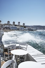 A coastal scene with several traditional windmills lined up on a hillside overlooking the sea. In the foreground, white wooden tables and chairs are arranged along a waterfront walkway, with waves crashing against the shore and splashing water into the air. The buildings have a white, Cycladic architectural style, complementing the clear blue sky and sea.