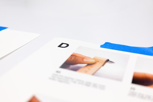 A close-up view of a printed page with a large letter 'D' and small images next to it, possibly illustrating steps or examples. The page is taped to a surface with a piece of blue tape, and there is a glimpse of another page partly visible on the left.