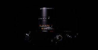 A close-up of a sleek camera lens partially illuminated, set against a dark background. The lens numbers and markings are visible, showcasing precision and detail.
