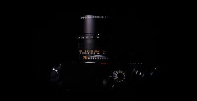 A close-up of a sleek camera lens partially illuminated, set against a dark background. The lens numbers and markings are visible, showcasing precision and detail.