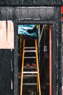 A person wearing work boots is standing on a yellow ladder inside a partially constructed or renovated building. The ladder is positioned in a doorway surrounded by dark, textured walls. Above the ladder is a blue tarpaulin covering something.