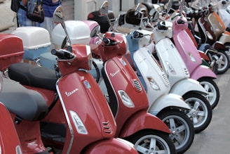 A lineup of colorful scooters parked closely together, featuring various shades including red, white, and pink. The scooters are equipped with rearview mirrors and some have storage compartments attached to the back. The scene suggests an urban setting.