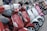 A lineup of colorful scooters parked closely together, featuring various shades including red, white, and pink. The scooters are equipped with rearview mirrors and some have storage compartments attached to the back. The scene suggests an urban setting.