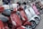 A lineup of colorful scooters parked closely together, featuring various shades including red, white, and pink. The scooters are equipped with rearview mirrors and some have storage compartments attached to the back. The scene suggests an urban setting.